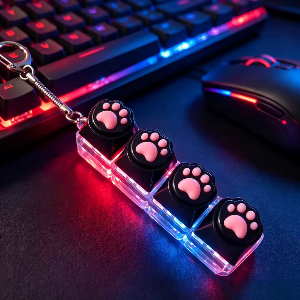 A keychain accessory featuring four transparent square blocks with black paw-print caps and pink paw-pad centers, resting on a desk near a keyboard and mouse with RGB lighting.