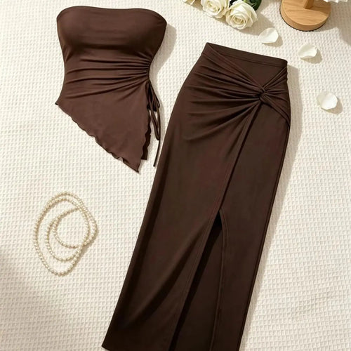 A matching two-piece women's outfit in dark brown, featuring a strapless ruched bandeau top with side ties and a high-waisted ruched midi skirt with a front slit, styled on a textured cream fabric with white roses and a pearl necklace.